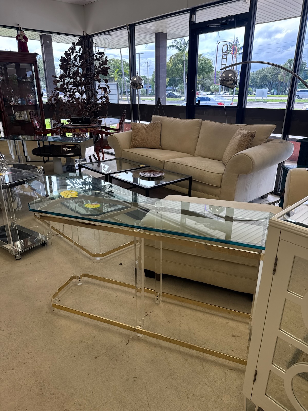 Showroom corner by the front windows with a cream rolled-arm sofa, a glass and lucite console table with brass trim, nesting glass coffee tables, a chrome arc lamp and a china cabinet