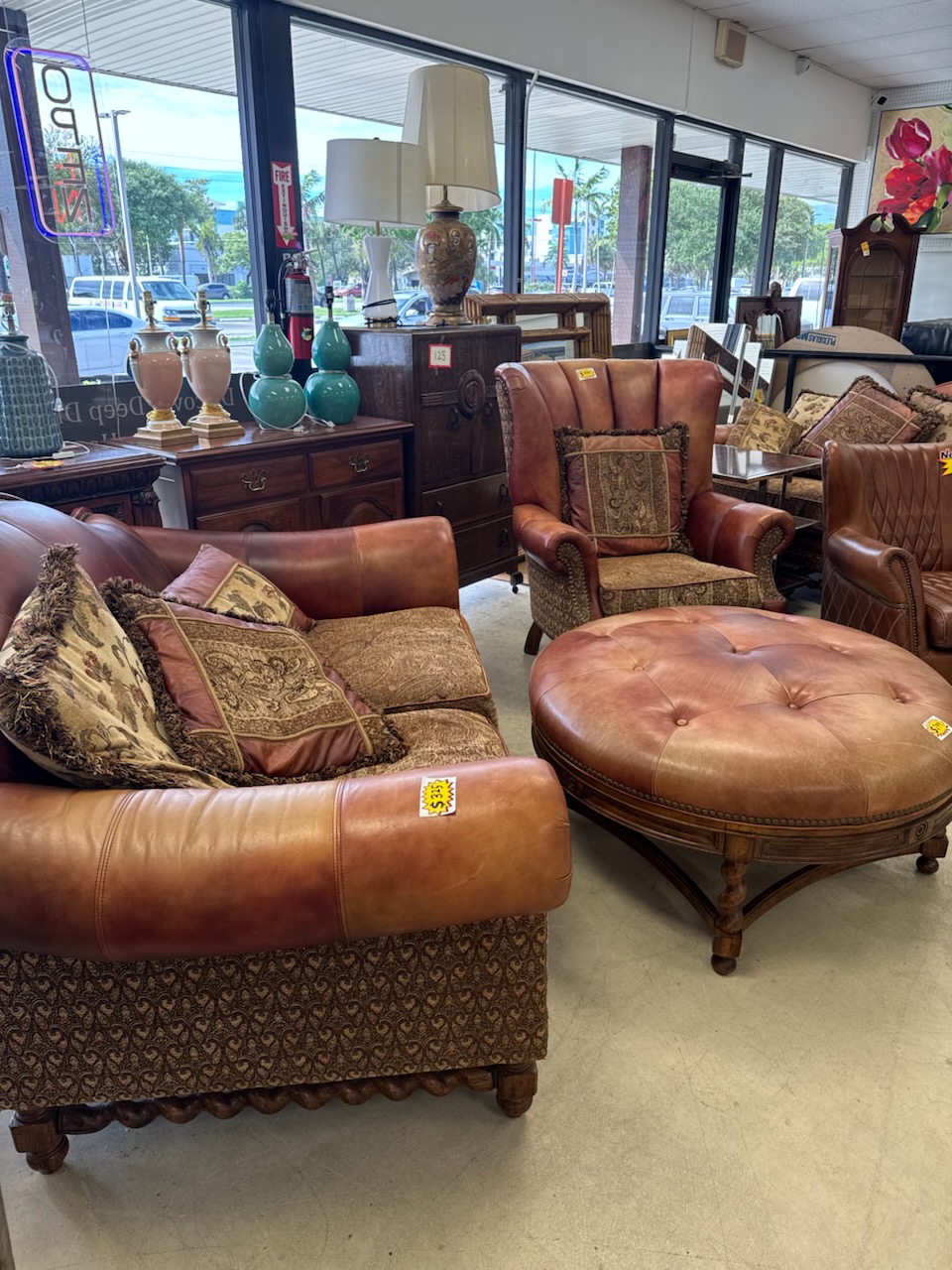 Brown leather and tapestry armchairs with fringed pillows around a large tufted leather ottoman, with table lamps, turquoise vases and wooden dressers along the windows