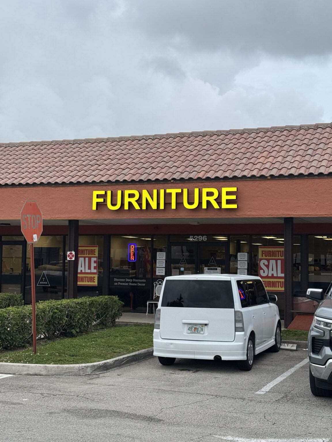 Storefront at 5296 N University Dr: a terracotta tile roof with a large yellow FURNITURE sign above glass doors marked 5296, with an OPEN sign and furniture sale posters in the windows